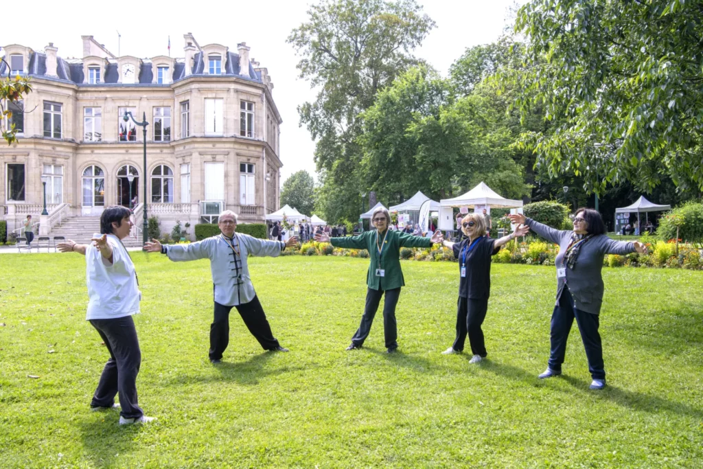 Des personnes âgées pratiquent le Tai Chi dans un parc devant un bâtiment historique.